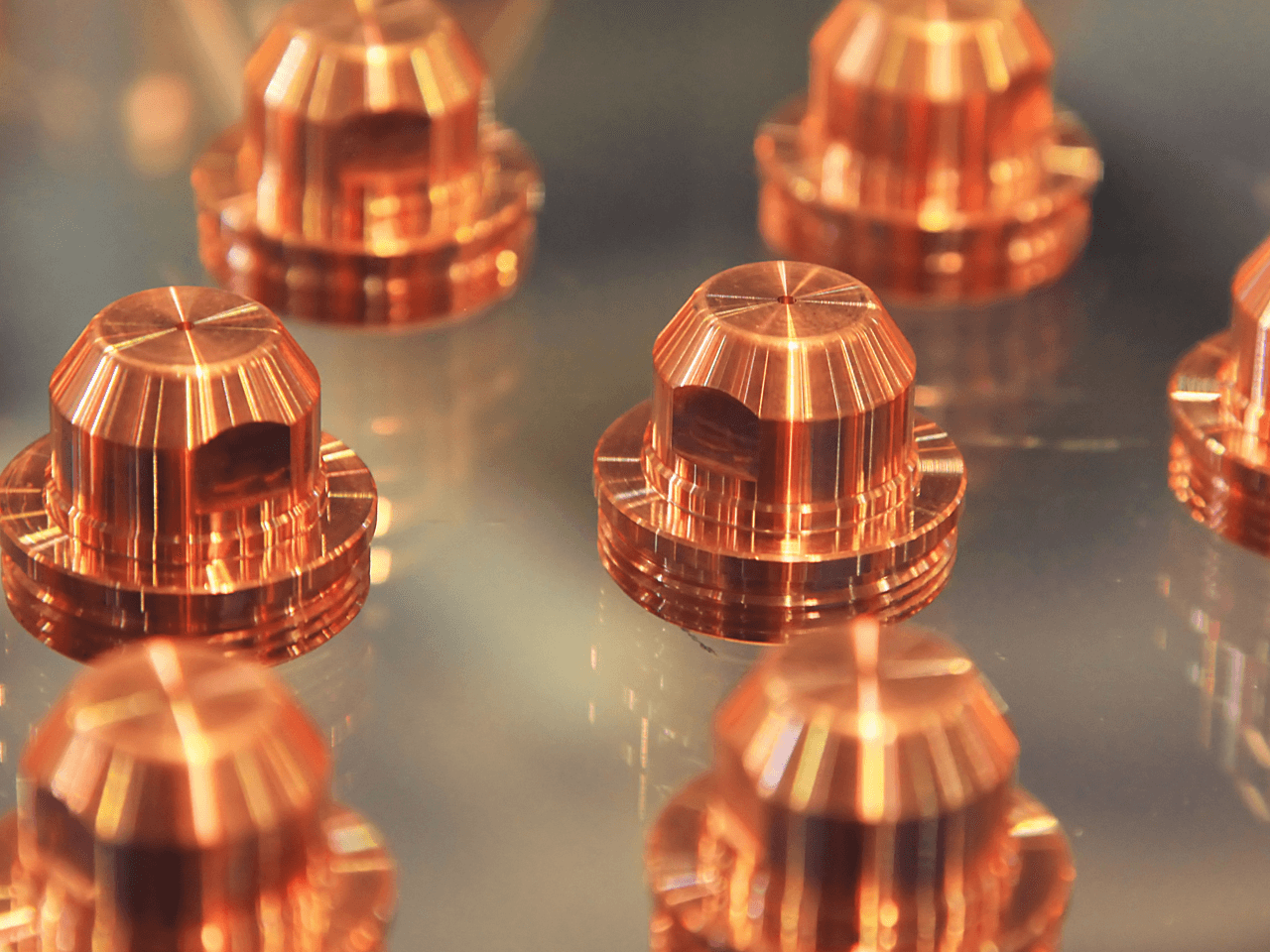 Copper-colored, cylindrical components with fluted tops are arranged on a reflective surface, their polished finish gleaming.