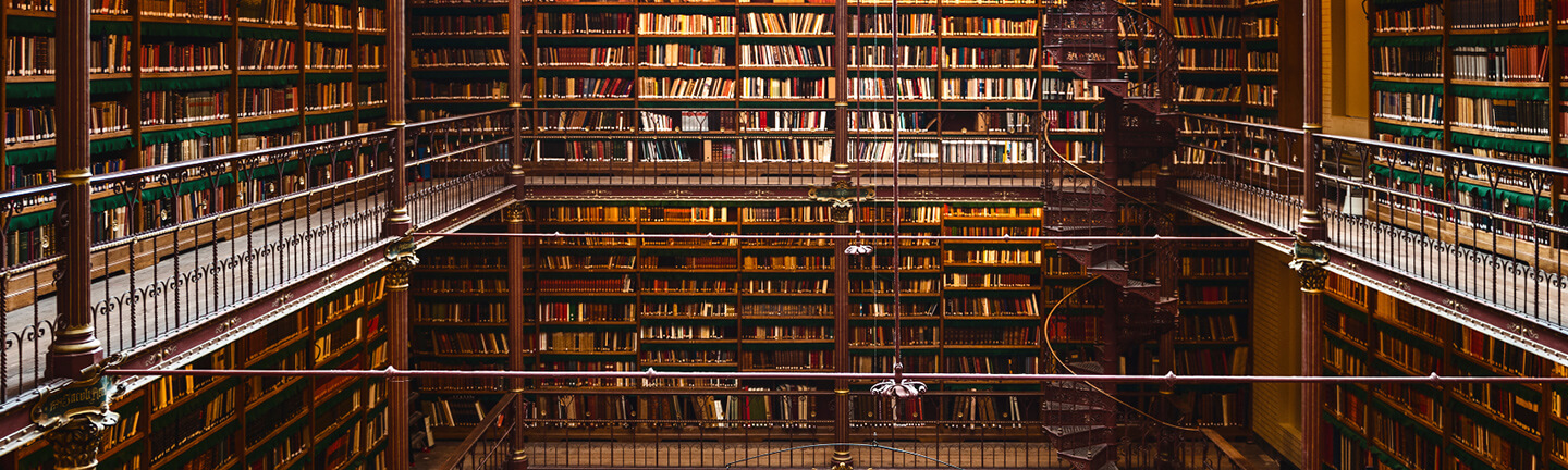 Books fill towering shelves in a large library with a spiral staircase and ornate metal railings.