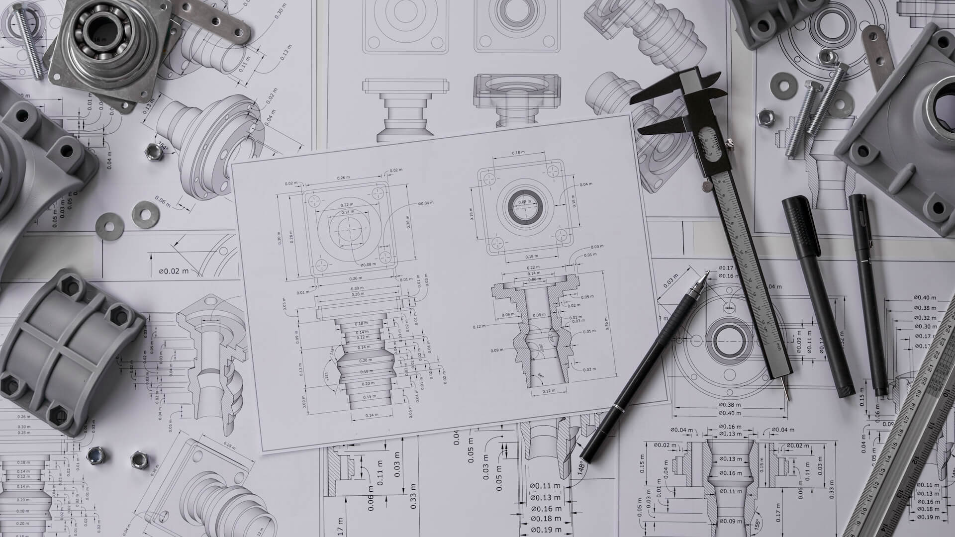 Mechanical components rest on detailed technical drawings with measurements, while calipers and pencils are placed nearby.