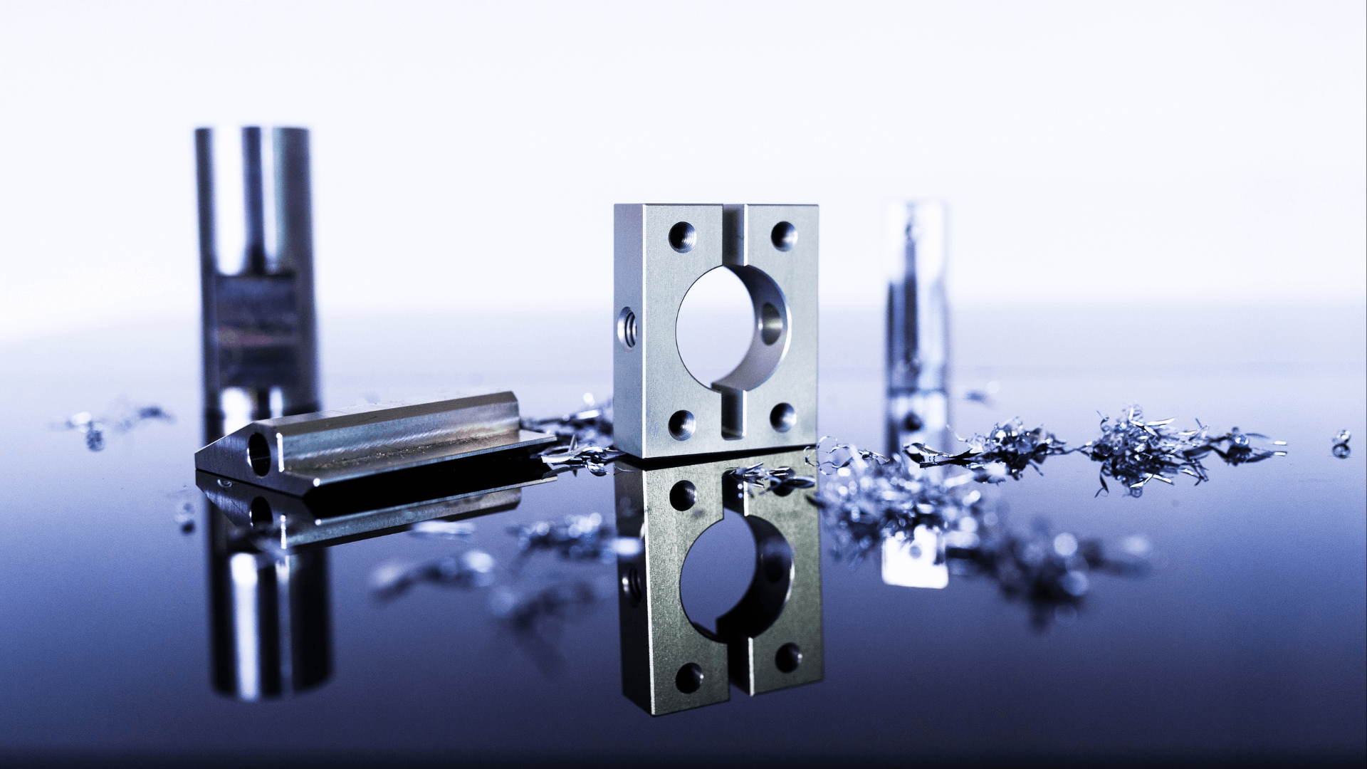 A metal bracket with a central hole stands upright on a reflective surface, surrounded by metal shavings & cylindrical parts.