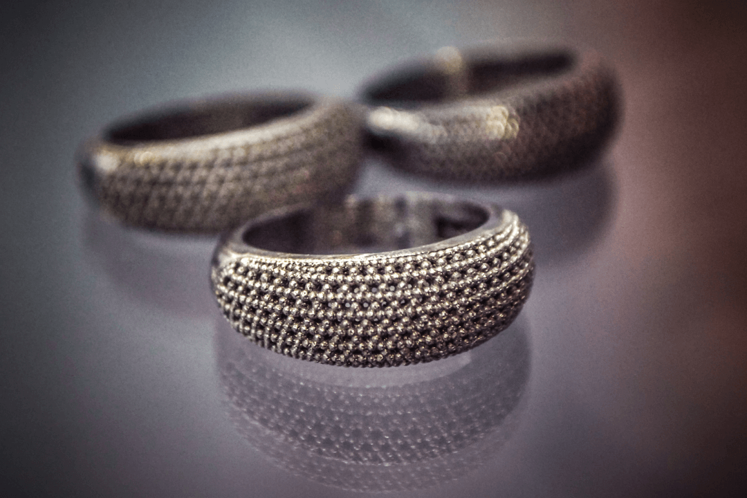 Three textured, detailed silver rings rest on a reflective surface, creating a soft reflection.