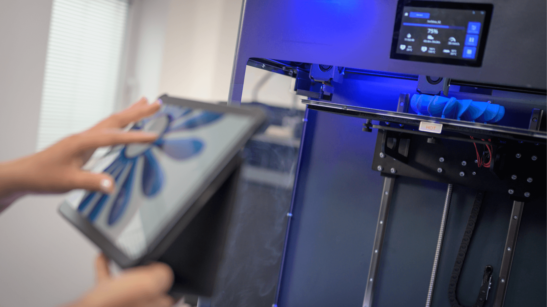 A 3D printer operates beside a person using a tablet displaying a blue fan design. A display above shows "75%".