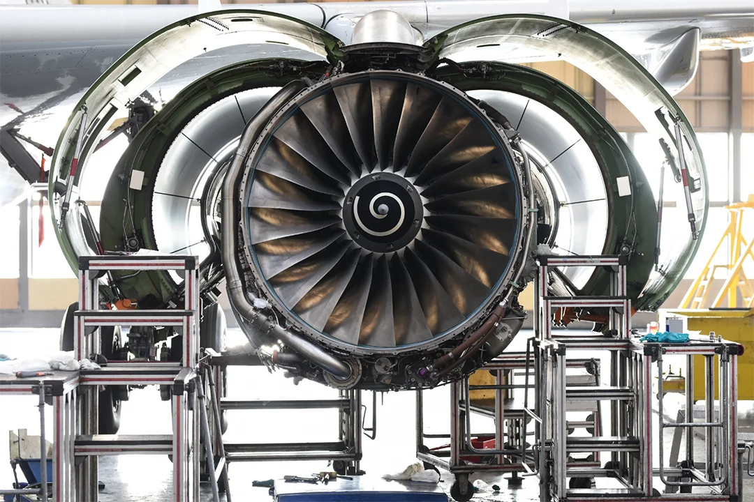 A jet engine is undergoing maintenance in a hangar, shown with its fan blades exposed and surrounded by scaffolding and tools.