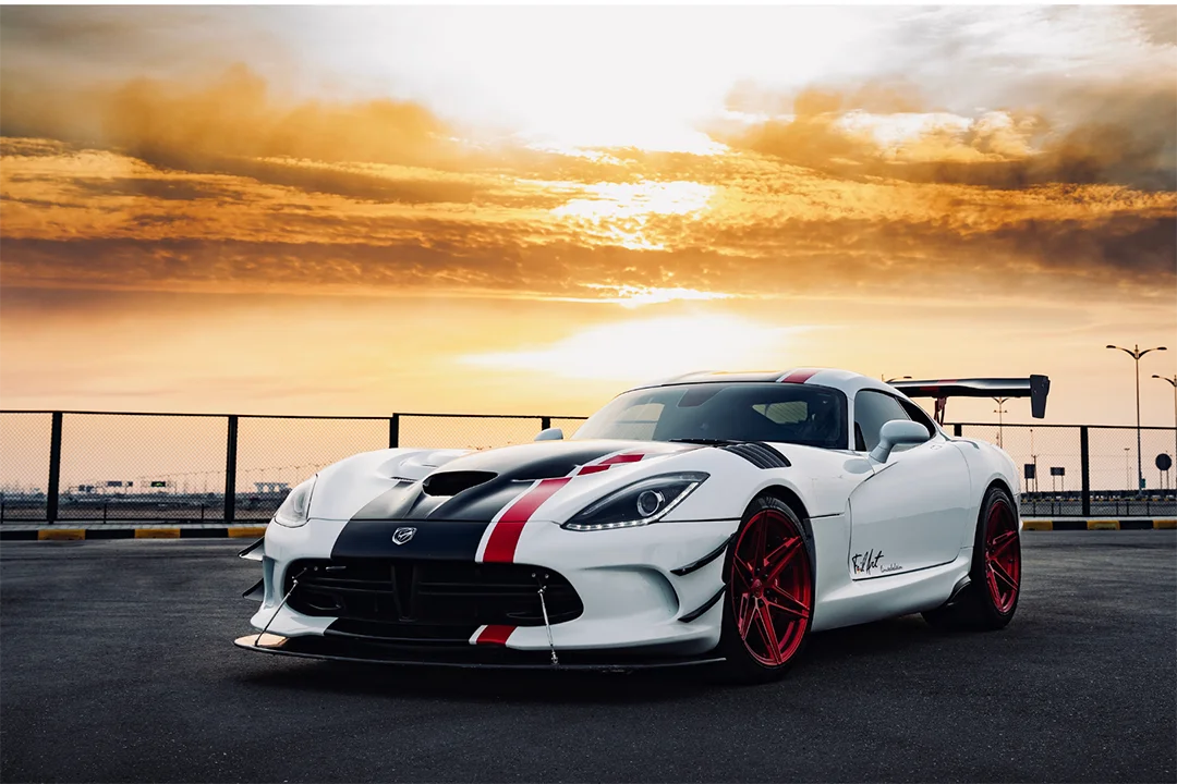 A white performance motorsports car with red stripes & rims is parked on a tarmac under a dramatic orange sunset sky, near a wire fence.