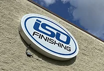 Close up of ISO Finishing logoed sign with blue letters against a white background on a beige building with blue sky and clouds in the background.