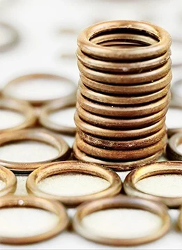 A stack of copper-colored rubber o-rings stacked upright with visible material flash from manufacturing, surrounded by additional o-rings scattered on a light-colored surface.