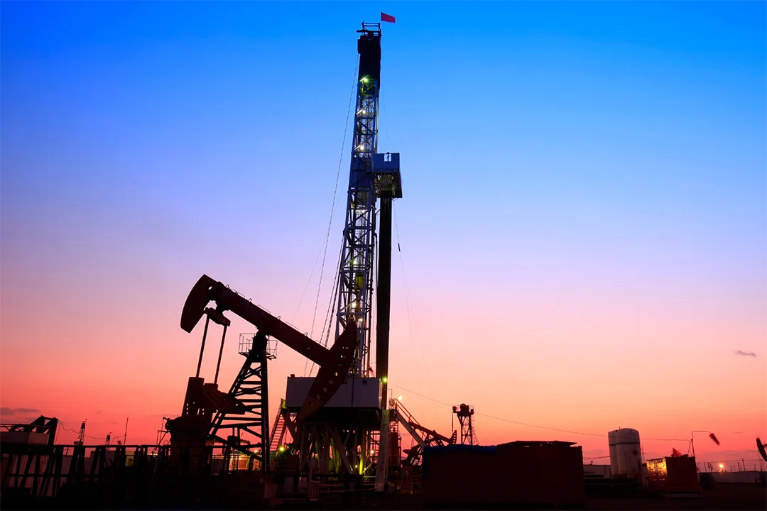 An oil drilling rig, surrounded by pump jacks, set against a colorful sunset sky.