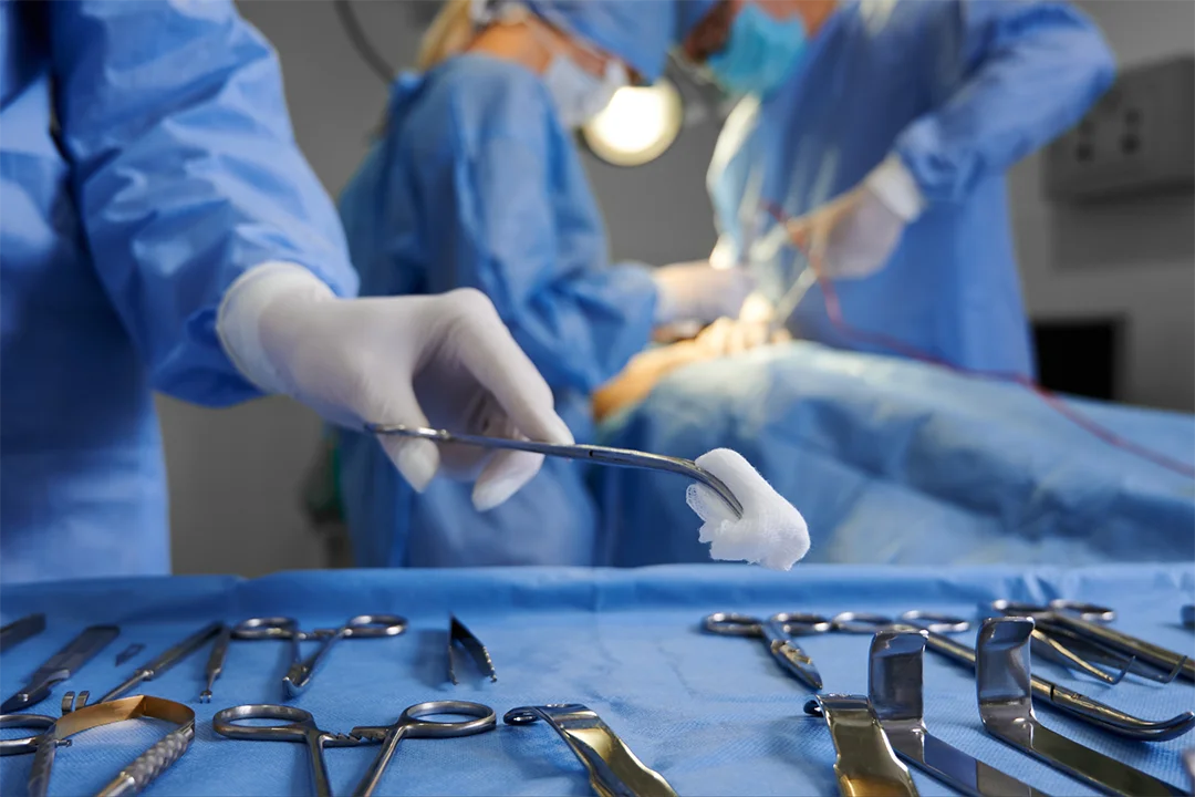 A surgeon holds gauze with forceps above a surgical instrument table, while others perform surgery in the background.