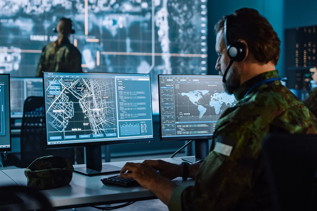 A person sits in a command center monitoring multiple computer screens displaying maps and data.