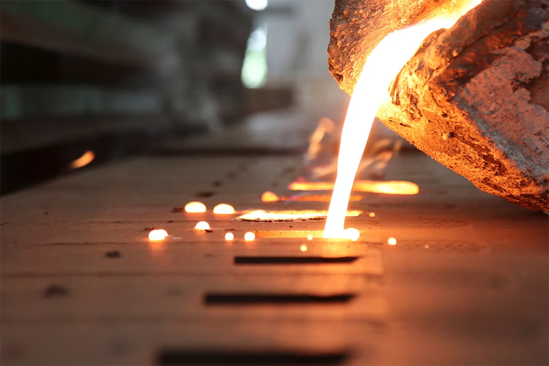 Molten metal flows from a crucible into a mold within a dim industrial setting with blurred shelves in the background.