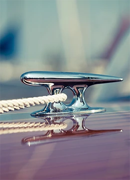 A polished, reflective metal cleat secures a rope on a wooden boat deck. The blurry background suggests a nautical setting.